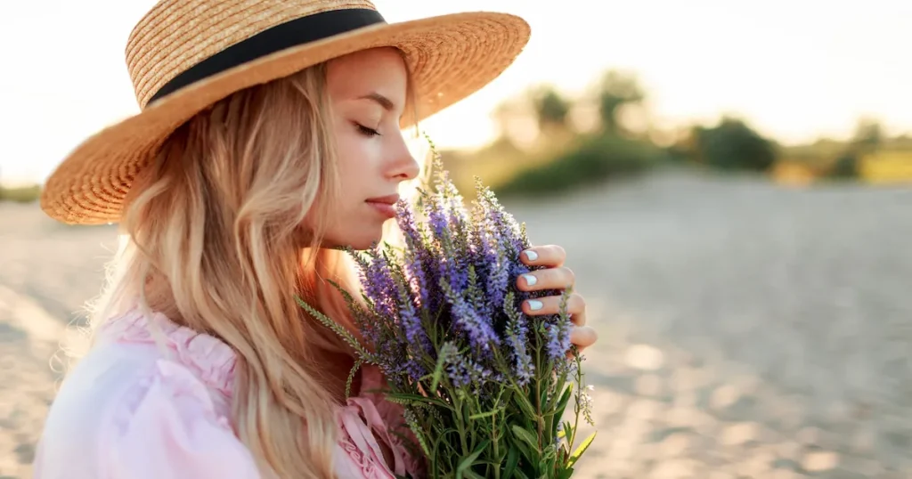 Romantic close up portrait of charming blonde girl in straw hat smells flowers on the evening beach, Warm sunset colors. Bouquet of lavender. Leef Sy Liefde prakties uit